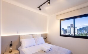 A modern bedroom with a neatly made bed, a nightstand with a lamp, and a window offering a city view. - 2BR apt w/ Grill 600m From the Beach Vre102 (Bombinhas)