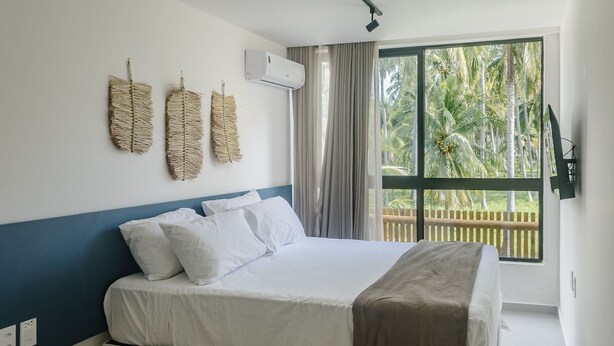 Un dormitorio con cama, almohadas blancas, una pared de acento azul, una ventana con vista a palmeras y un aire acondicionado instalado en la pared.