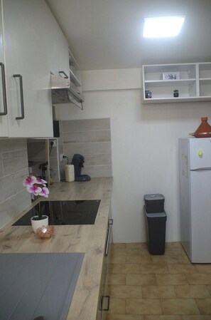 A kitchen with white cabinets, a black countertop, a white refrigerator, a trash bin, and a small shelf with decorative items. - Typical Corsican apartment (Cervione)