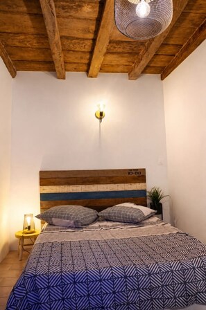 A bedroom with a wooden headboard, a bed with patterned bedding, a wall-mounted light fixture, and a small table with a lamp. - Typical Corsican apartment (Cervione)