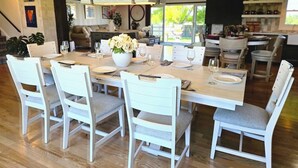 Zona pranzo con un grande tavolo in legno apparecchiato per sei persone, circondato da sedie bianche con cuscini grigi.