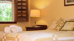 A neatly made bed with a quilt, a bedside lamp, rolled towels, and a window with shutters. - Queen-size bed, with a private bathroom, a walk-in shower and closet. (Orchid Bay)