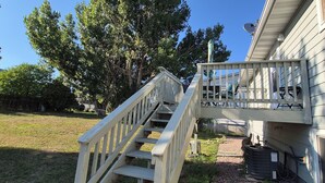 Un escalier en bois mène à un espace extérieur abrité, doté d’une rambarde en métal, et entouré d’arbres et d’une pelouse.