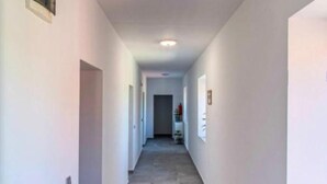 Un pasillo estrecho y bien iluminado, con paredes blancas y alfombra gris. Hay puertas a ambos lados y un pequeño estante con objetos en la pared derecha.