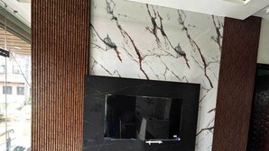 A modern living room with a large TV mounted on a marble wall, wooden paneling, and a wooden console table. - Hotel Moad Mudgal Palace (Khandwa)
