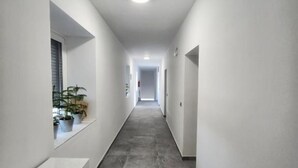 A narrow, well-lit hallway with a tiled floor and white walls. There are potted plants on the left side and a door at the end of the hallway. - Rooms Burnum - Twin Room 7 (Vrbnik-Knin)