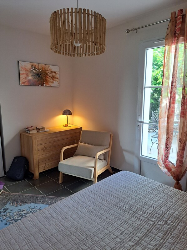 A bedroom with a bed, a wooden dresser, a chair, a lamp, and a window with curtains. - This charming house, rated 3* by S'I de Royan, features a swimming pool (Vaux-sur-Mer)