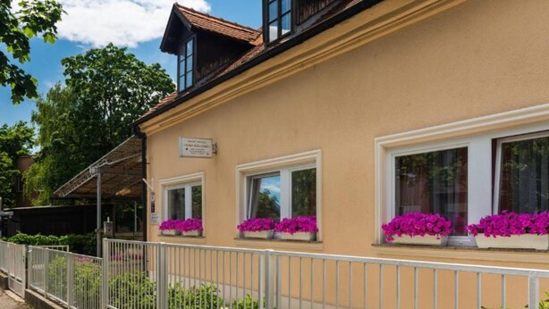 Ein Gebäude mit einer beige gefärbten Fassade, mehreren Fenstern mit Blumenkästen und einem weißen Metallzaun.
