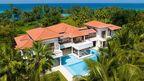 A large, modern house with a swimming pool, surrounded by lush greenery and palm trees, overlooking a clear blue ocean.
