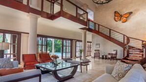 A spacious living area with a glass coffee table, comfortable seating, a staircase, and a large window offering a view of the outdoors.