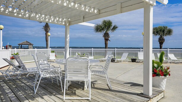 Un espace extérieur aménagé, abrité et doté de chaises et d’une table en métal blanc, offrant une vue sur une plage bordée de palmiers et délimitée par une clôture blanche.