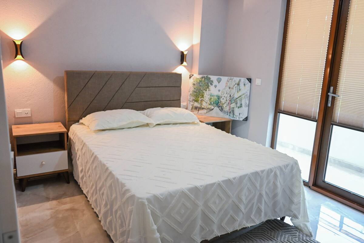 Un lit bien fait, recouvert d’une couvre-lit blanche, deux tables de chevet avec des lampes, une œuvre d’art accrochée au mur et une fenêtre avec des stores.