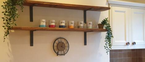 A kitchen with a wooden shelf holding mugs, a wall clock, and a bowl on the counter.