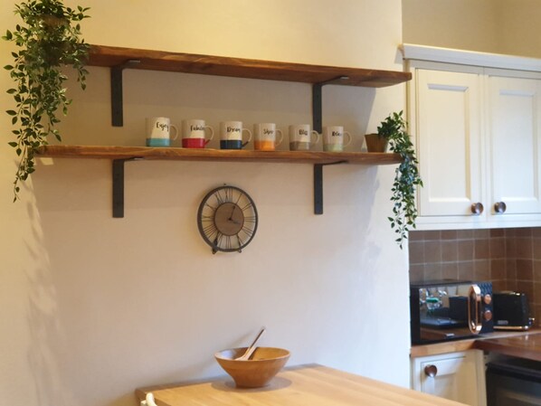A kitchen with a wooden shelf holding mugs, a wall clock, and a bowl on the counter.