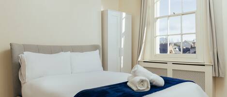 A neatly made bed with white and blue bedding, a window with a view of buildings, and a white wardrobe.