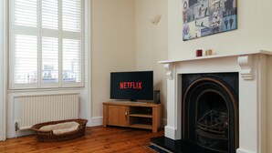 Ein Wohnzimmer mit Kamin, einem Fernseher, der 'NETFLIX' anzeigt, einem Korbwägelchen und einer Heizung.