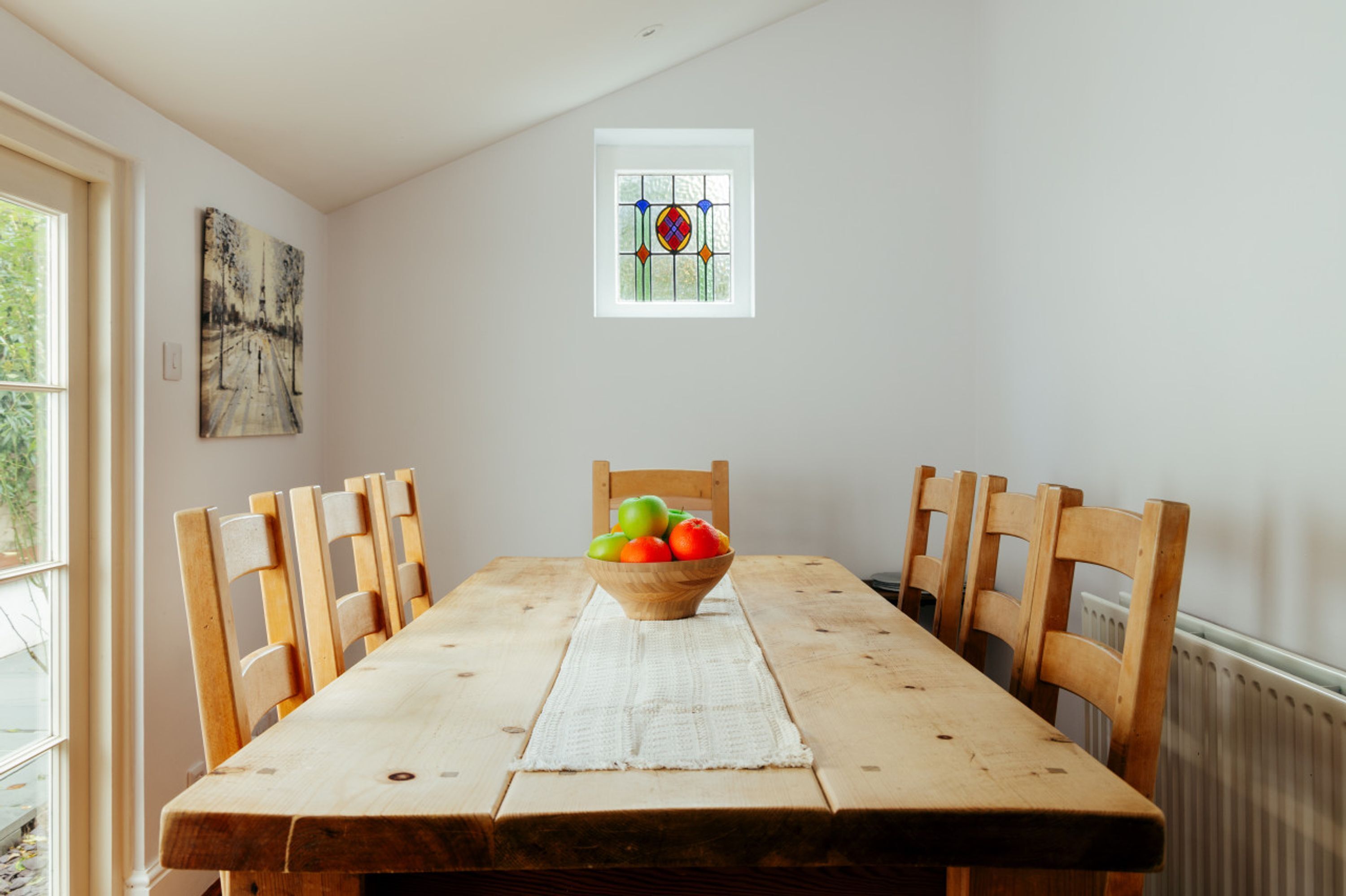 A dining table with a bowl of fruit, wooden chairs, a white table runner, a painting, and a window with a stained glass design.