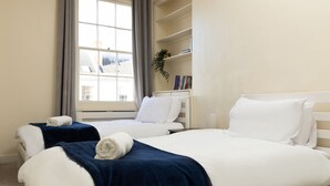Ein Hotelzimmer mit zwei Betten, einem Fenster mit Vorhängen, einem Bücherregal und einer Topfpflanze.