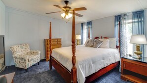 A four-poster bed with a wooden frame and a ceiling fan, a wooden nightstand with a lamp, a patterned armchair, and a wooden dresser.