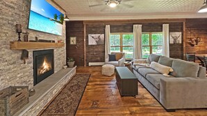 A cozy living room with a fireplace, a large TV, a ceiling fan, and a view of the outdoors through large windows. - Cozy Lake Cabin in Restored 1894 Schoolhouse—Perfect Escape. (Aitkin)
