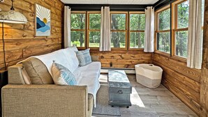 A cozy cabin interior with a wooden wall, a sofa, a small table, and a window with curtains. - Cozy Lake Cabin in Restored 1894 Schoolhouse—Perfect Escape. (Aitkin)