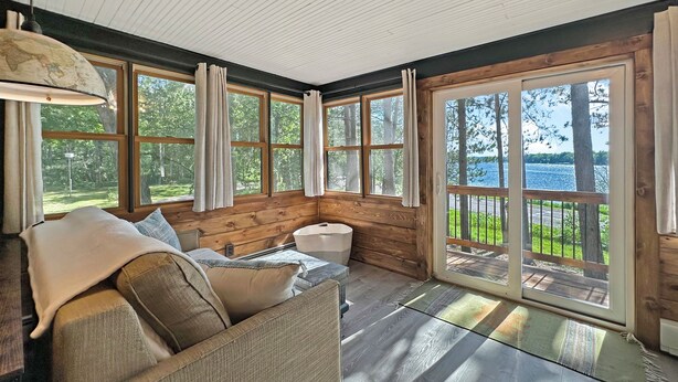 A cozy cabin interior with a wooden interior, a sofa with cushions, a large window with curtains, and a view of a lake. - Cozy Lake Cabin in Restored 1894 Schoolhouse—Perfect Escape. (Aitkin)