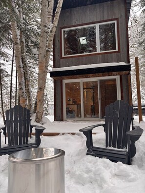 Una cabaña de madera de dos pisos con entrada por una puerta de cristal, rodeada de terreno cubierto de nieve y abedules.