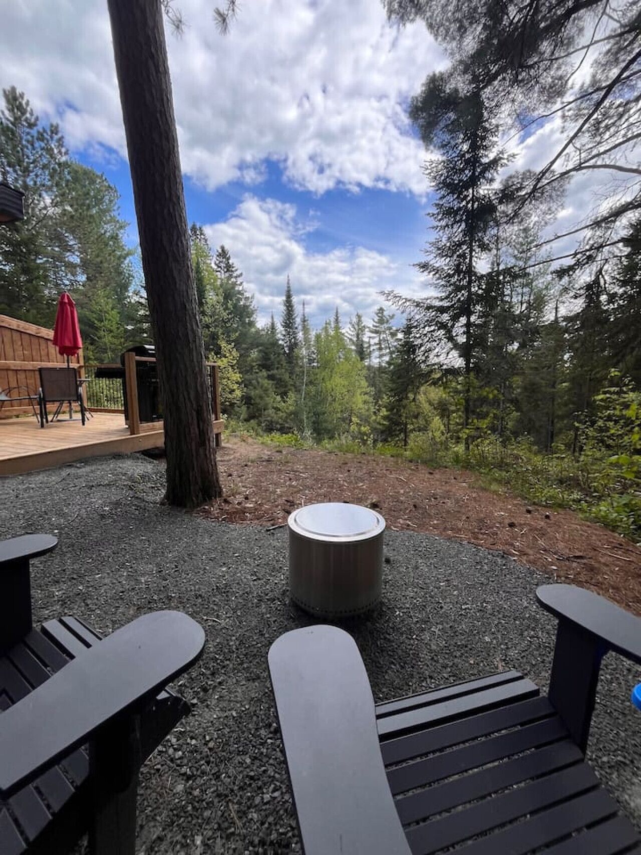 Un espace extérieur aménagé avec une table ronde et des bancs, entouré d’arbres et d’une terrasse en bois.