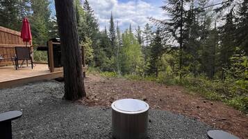 Un espace extérieur aménagé avec une table ronde et des bancs, entouré d’arbres et d’une terrasse en bois.
