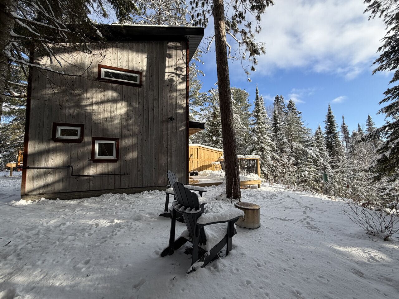 A wooden cabin with a snow-covered deck, a bench, and a chair.