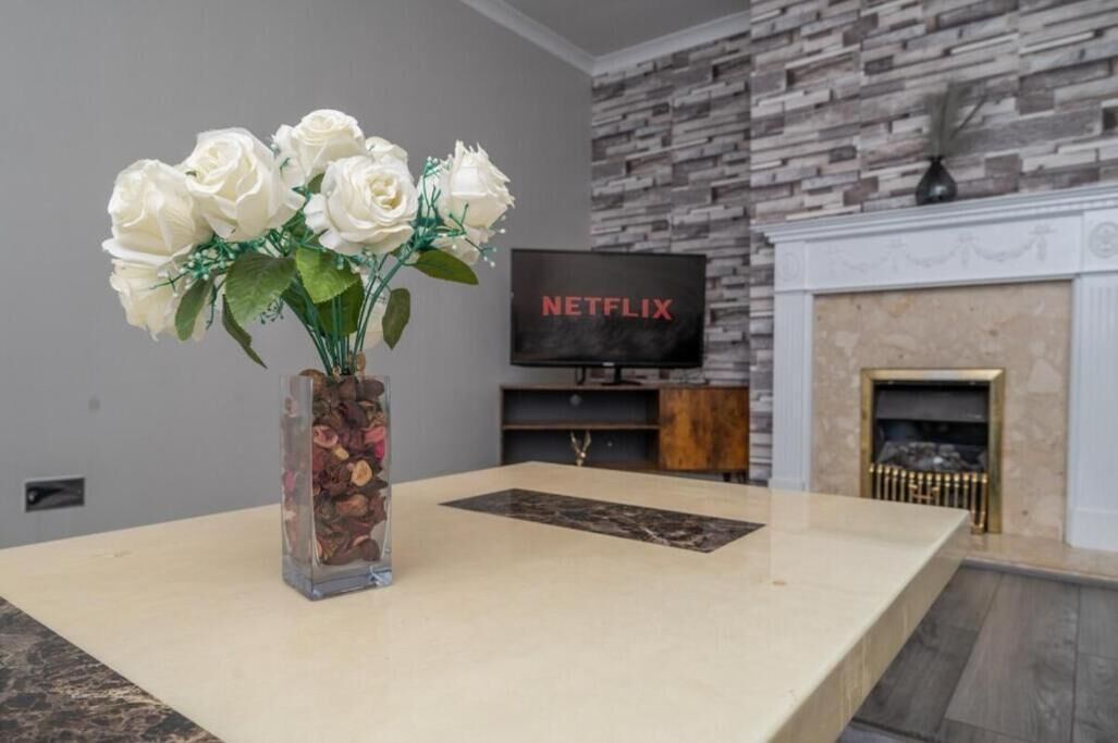 Una sala moderna con pared de piedra, chimenea, televisor y un florero con rosas blancas sobre una mesa.