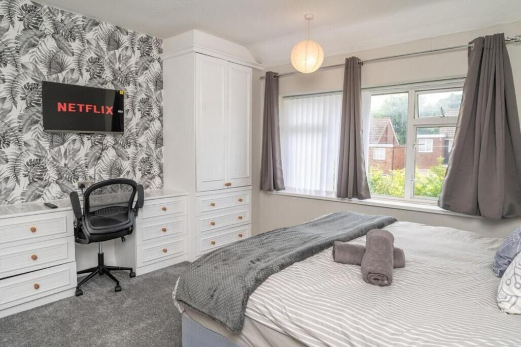 Ein Schlafzimmer mit einem Bett, einem Schreibtisch mit Stuhl, einem Kleiderschrank, einem an der Wand befestigten Fernseher und einem Fenster mit Vorhängen.