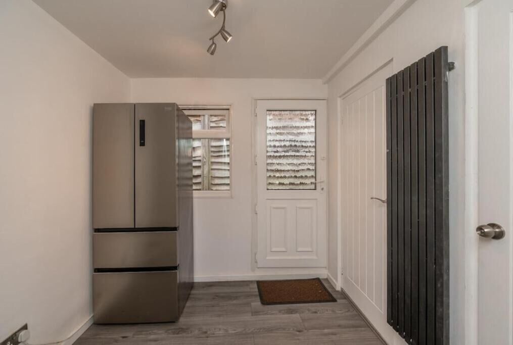 A narrow hallway with a stainless steel refrigerator, a white door with a window, and a radiator.