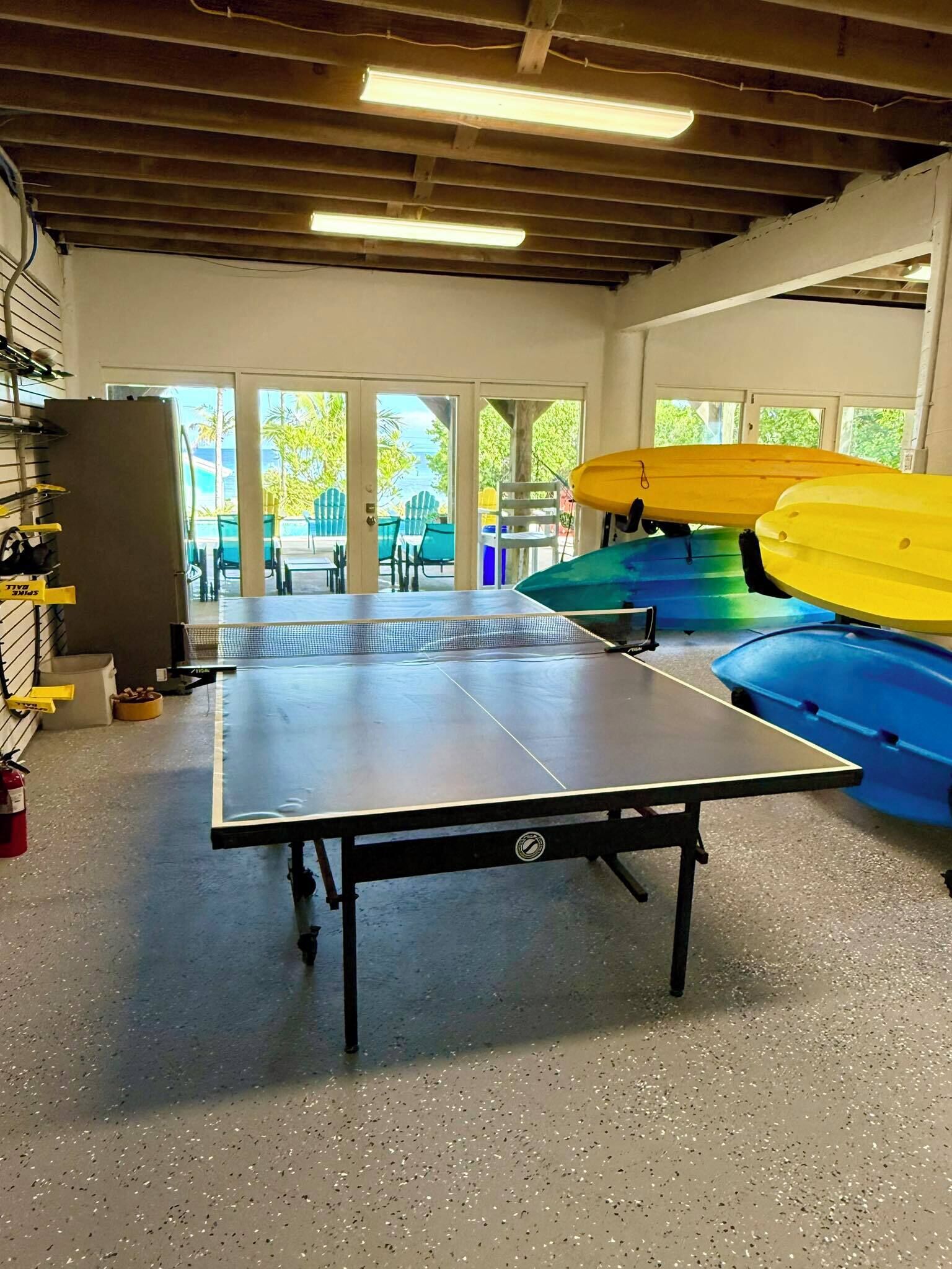A ping pong table in a room with a view of outdoor seating and kayaks stored nearby.