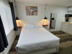 A neatly made bed in a hotel room with a bedside table, a wall-mounted mirror, and a small shelf with a lamp.