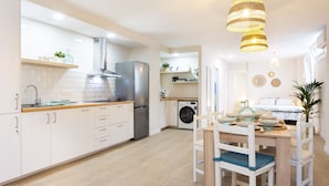 Una cocina moderna con armarios blancos, un refrigerador de acero inoxidable, un horno empotrado y una lavadora. Hay un comedor con una mesa y sillas de madera.