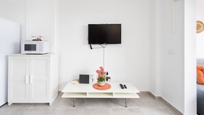 Un salon moderne avec un téléviseur à écran plat fixé au mur, un meuble blanc contenant un micro-ondes, une table basse blanche ornée d’un vase de fleurs et une télécommande.