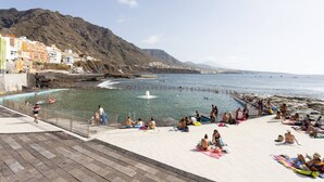 A coastal promenade with a swimming pool, beach, and mountainous backdrop.