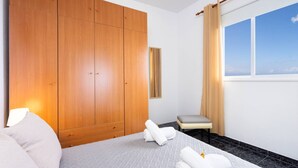 A hotel room with a large wooden wardrobe, a bed with white bedding, a small bedside table, and a view of the sky through a window with curtains.