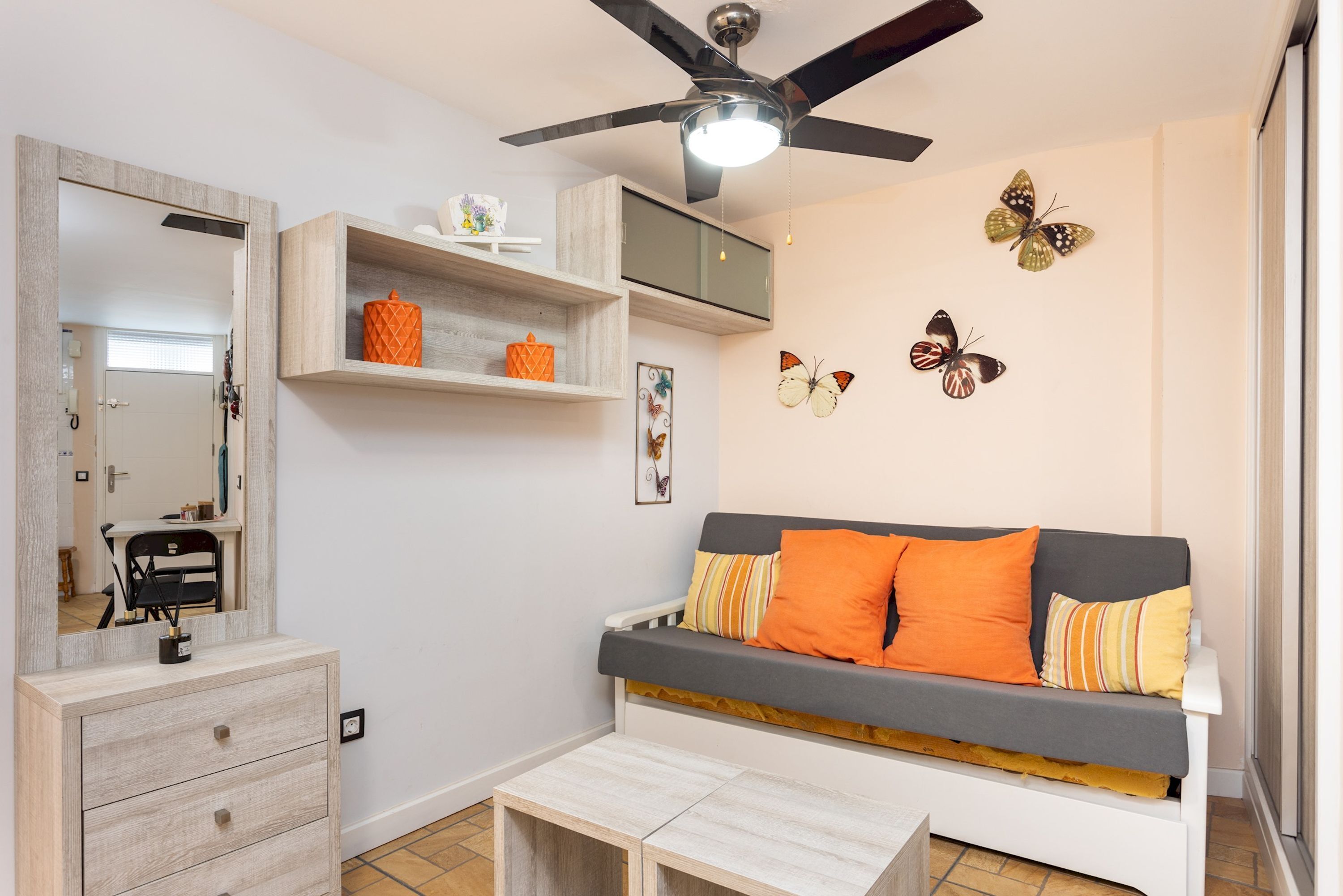 A modern living room with a grey sofa, orange cushions, a wooden coffee table, a ceiling fan, and butterfly wall art.