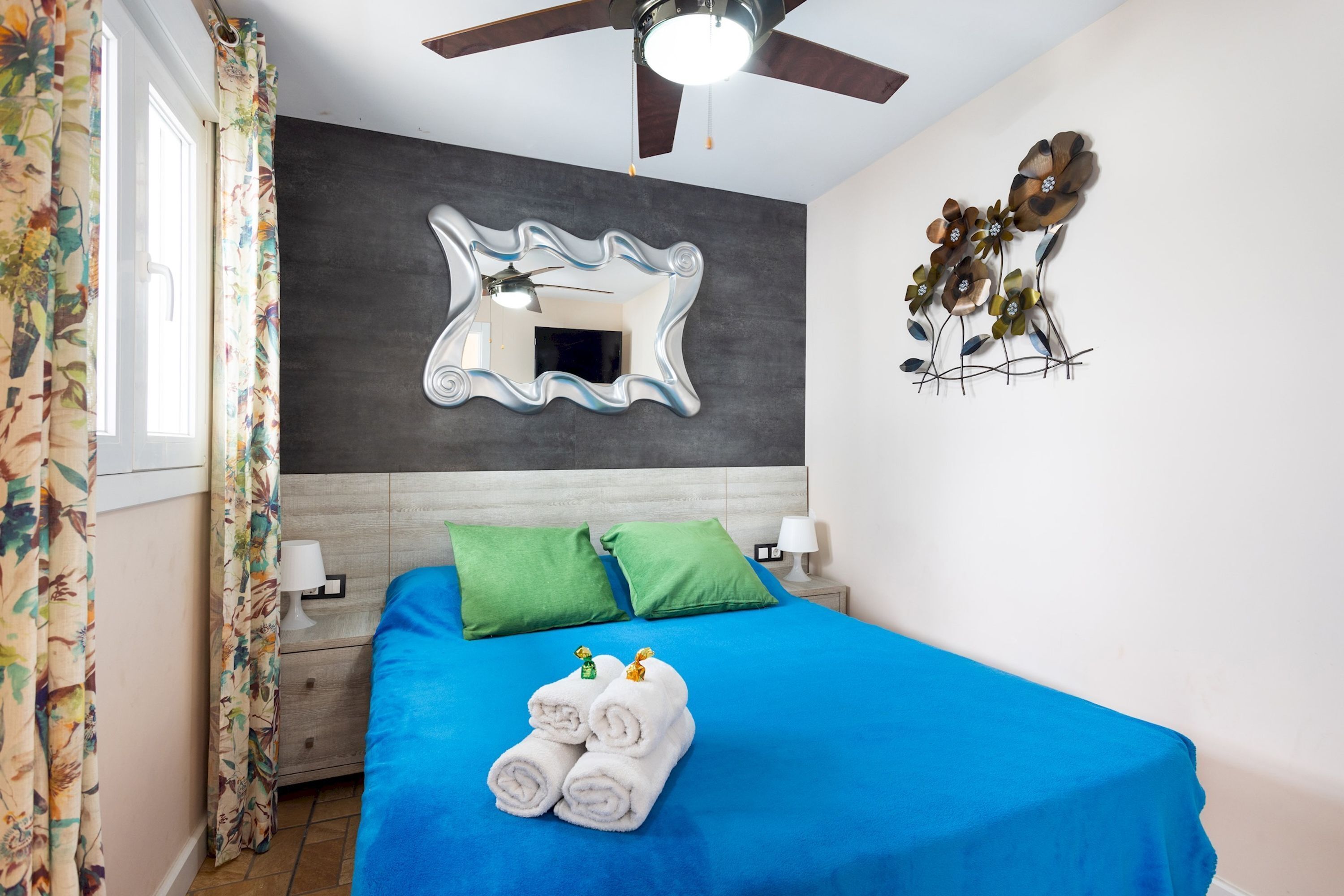 A bedroom with a blue bedspread, green pillows, a large mirror, and a ceiling fan.