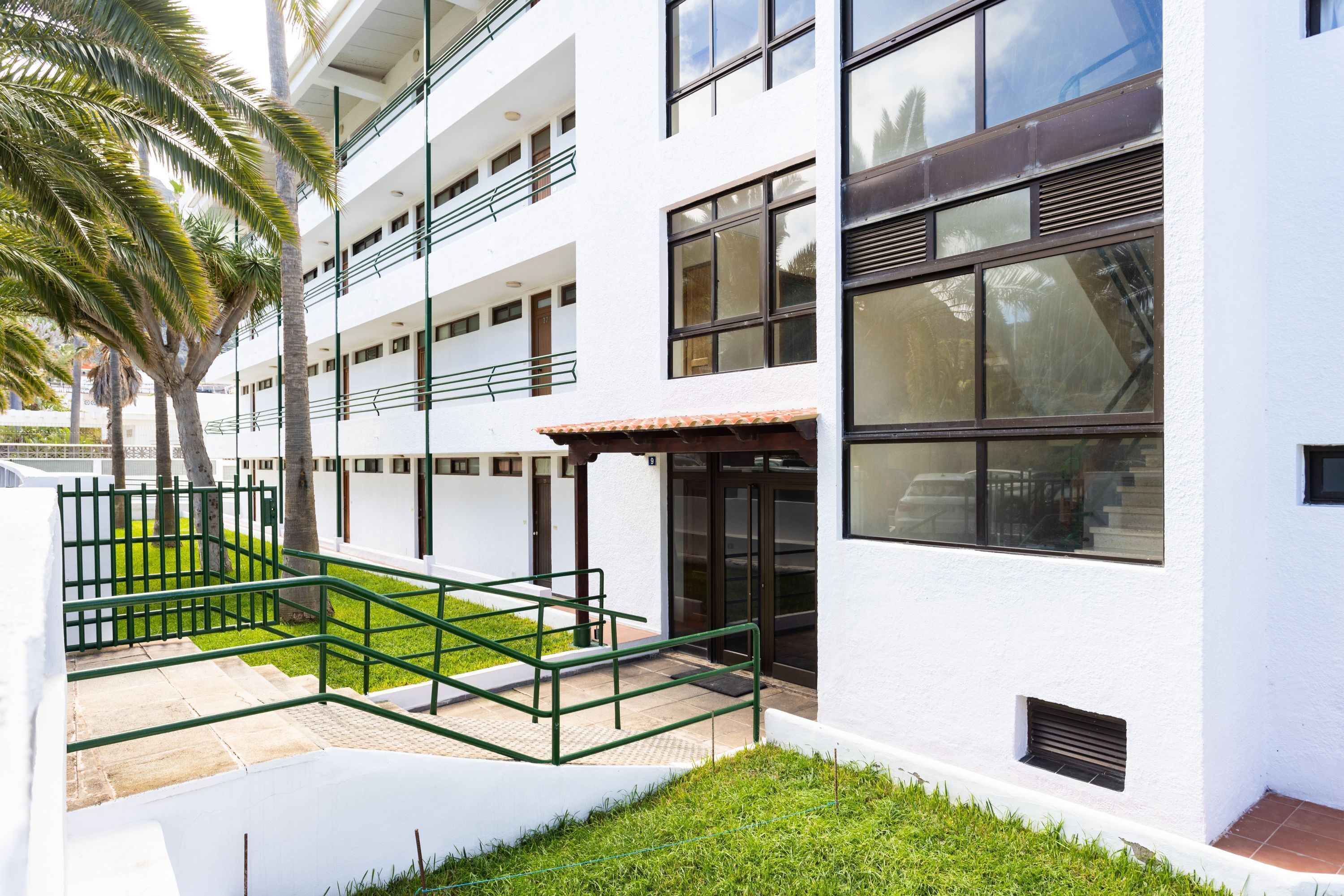 Un edificio moderno con varios pisos, amplias ventanas y un espacio exterior verde con escaleras y barandillas.