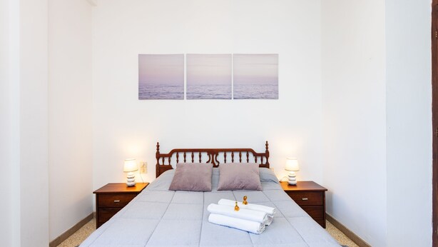 Un lit bien fait, recouvert d’une couvre-lit bleue, avec deux oreillers et une pile de serviettes pliées posée sur le lit. Il y a deux tables de chevet avec des lampes, et une œuvre d’art accrochée au mur au-dessus du lit.