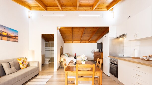 Een moderne keuken met houten kastjes, een eethoek met een houten tafel en stoelen, en een zithoek met een bank en een schilderij aan de muur.