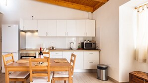 Een kleine keuken met witte kastjes, een houten eettafel met vier stoelen, een roestvrijstalen spoelbak, een magnetron en een prullenbak.