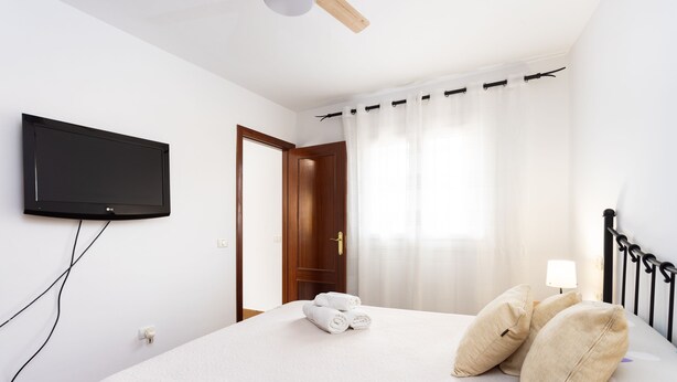 A neatly made bed with rolled towels, a television mounted on the wall, and a door leading to another room. - Piso Elegante y Luminoso (La Victoria de Acentejo)
