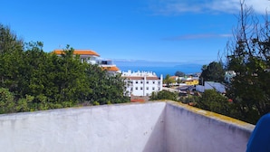 Un balcone con vista su una cittadina costiera, con edifici dai tetti arancioni e un cielo azzurro intenso.