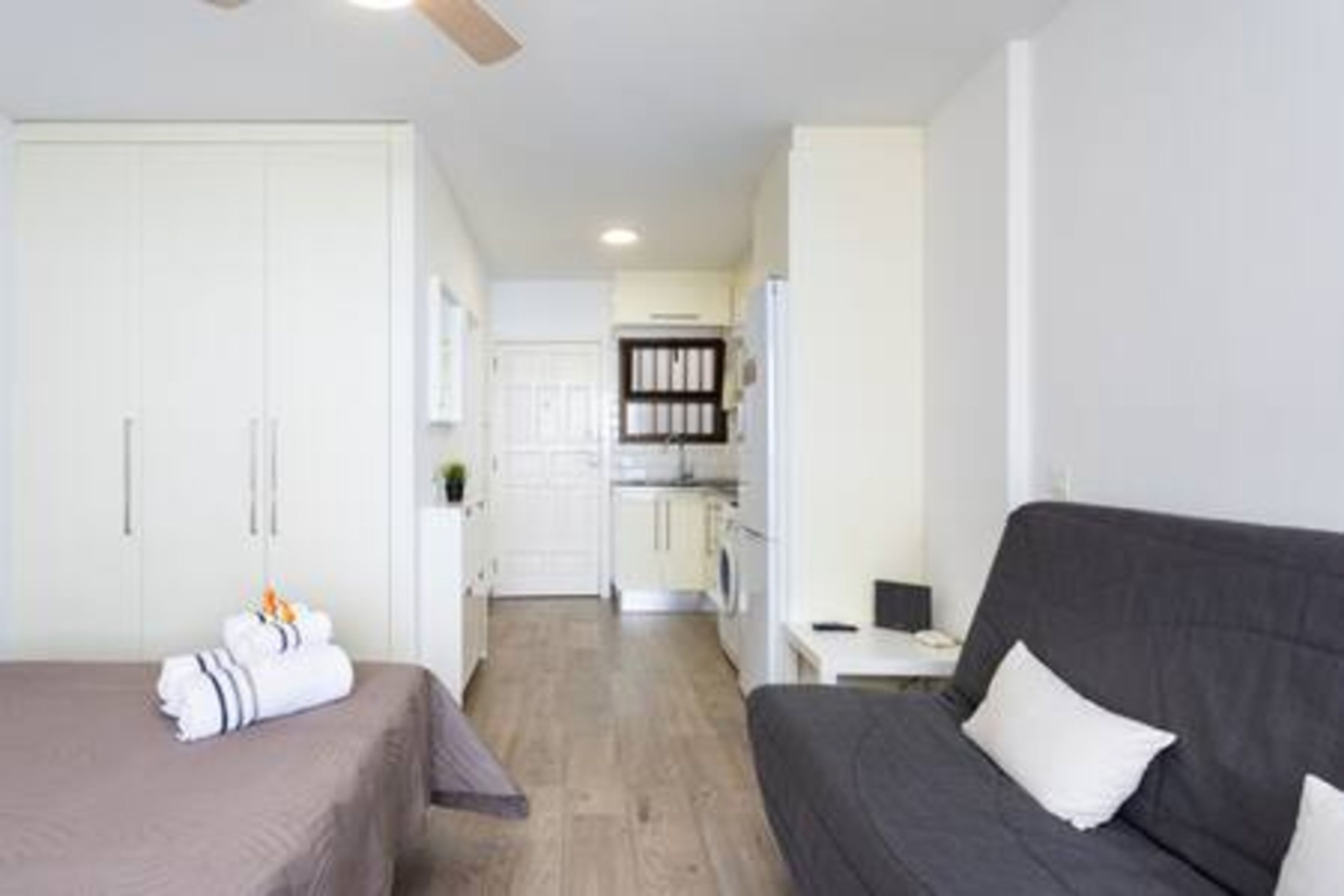 A modern hotel room with a bed, a sofa, a desk, and a kitchenette in the background.