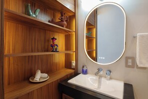 A bathroom with a wooden vanity, a white sink, a round mirror with a light, and shelves with decorative items. - Luxury Casita Calle 47 Gastronomic Corridor (Mérida)