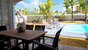 Un espace aménagé au bord de la piscine, avec des chaises longues et une table, entouré de palmiers et surplombé d’un ciel bleu azur.